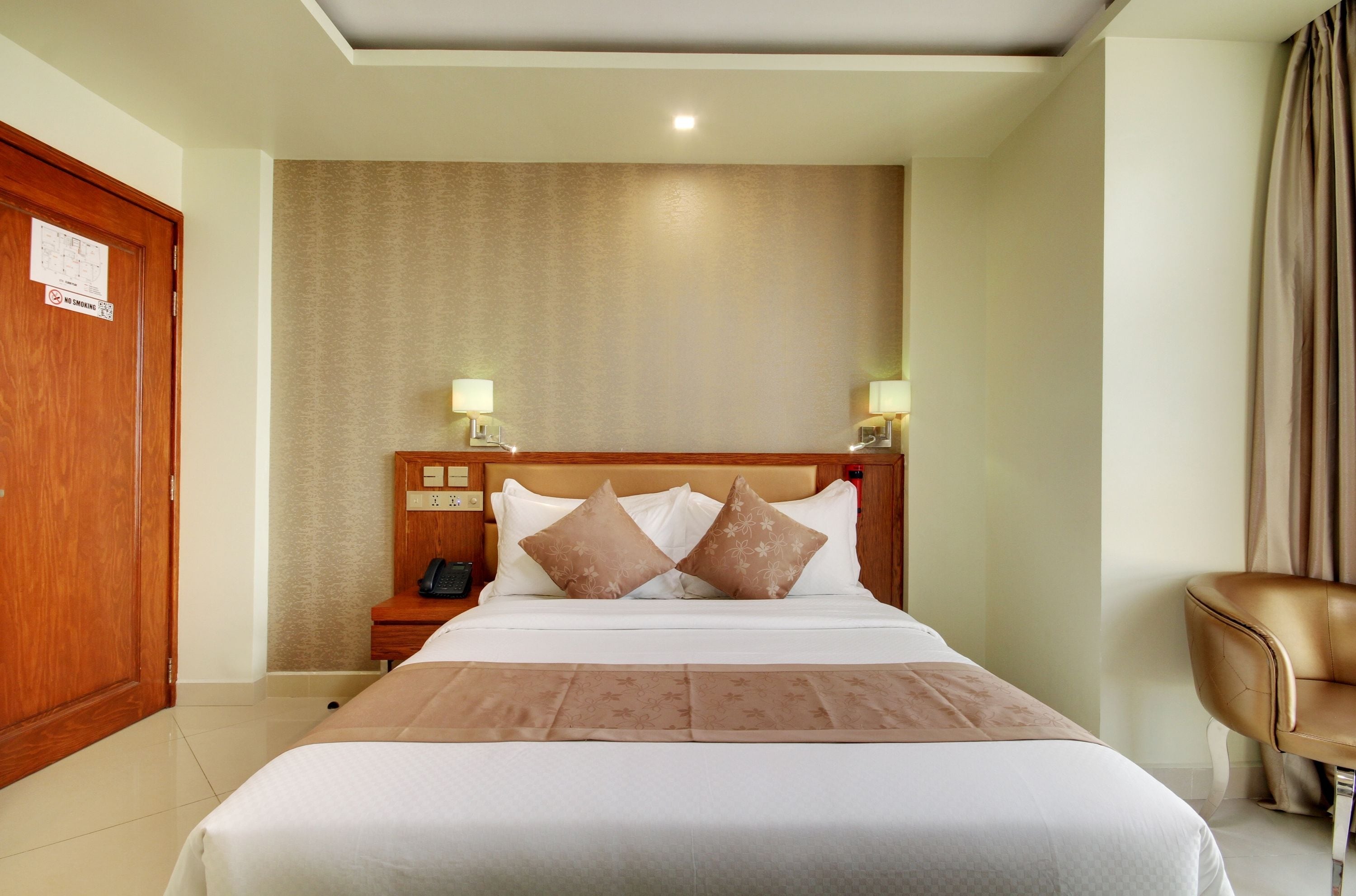 A hotel room with a large bed, a wooden headboard, a nightstand with a lamp, a chair, and a door with a no-smoking sign.