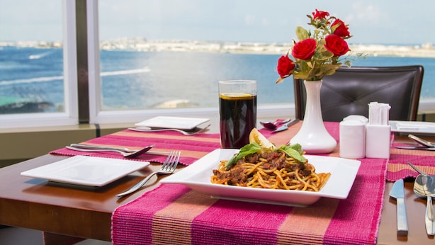 Una mesa de comedor con un plato de espagueti, un vaso de bebida oscura y un florero con rosas rojas.