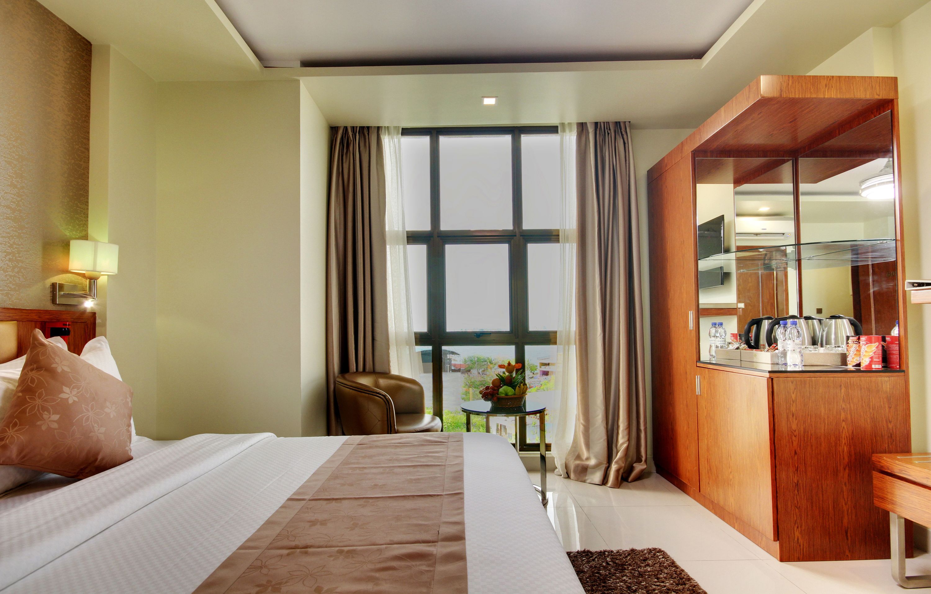 A hotel room with a bed, a desk, a chair, a small table with a plant, and a view of the outdoors through large windows.