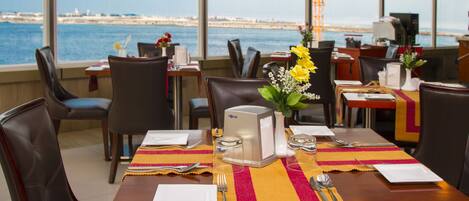 A restaurant with a view of the sea, tables set with yellow and red placemats, and a coffee machine on the table.