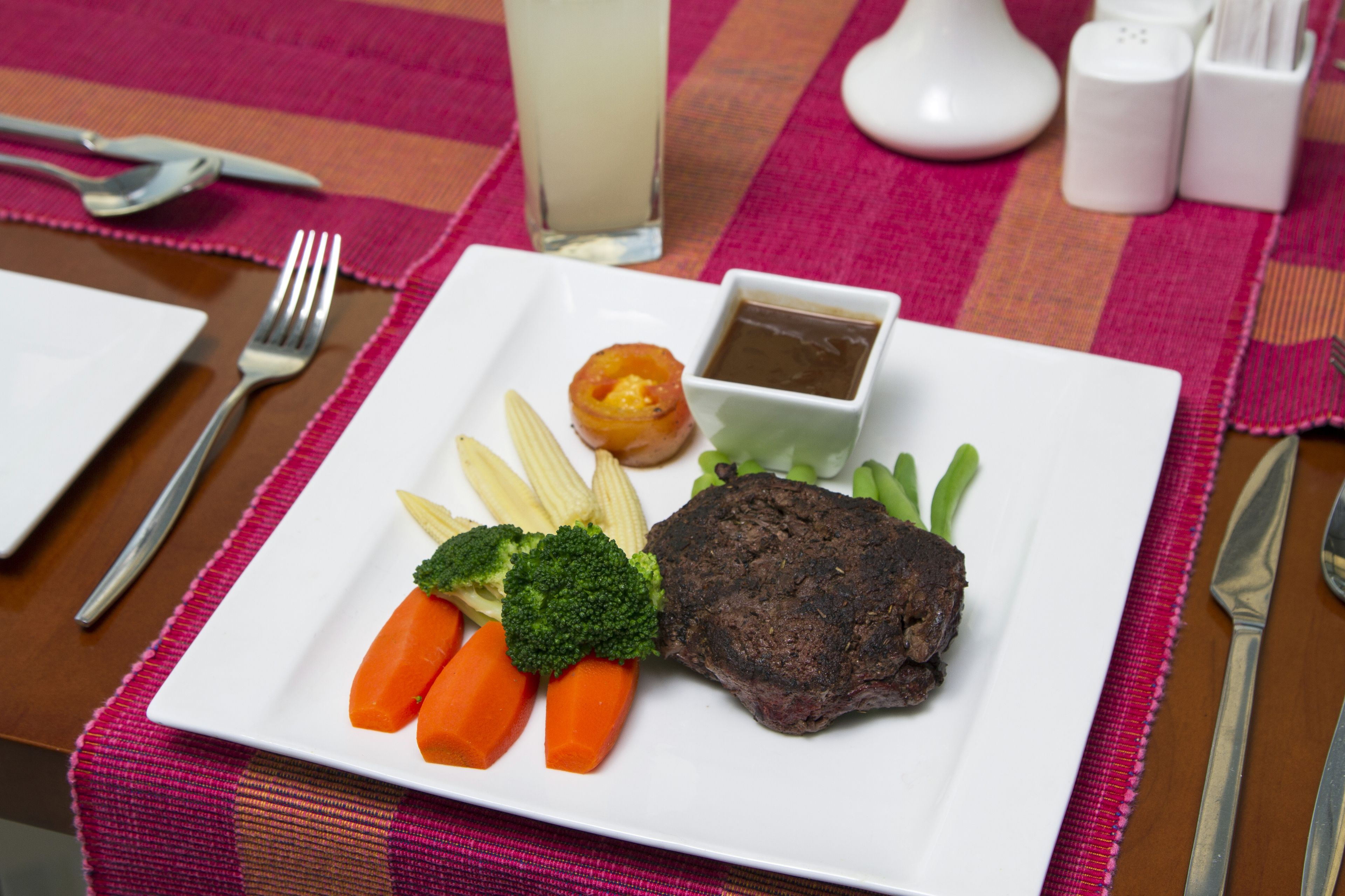 A plate with a steak, broccoli, carrots, and a small bowl of sauce.