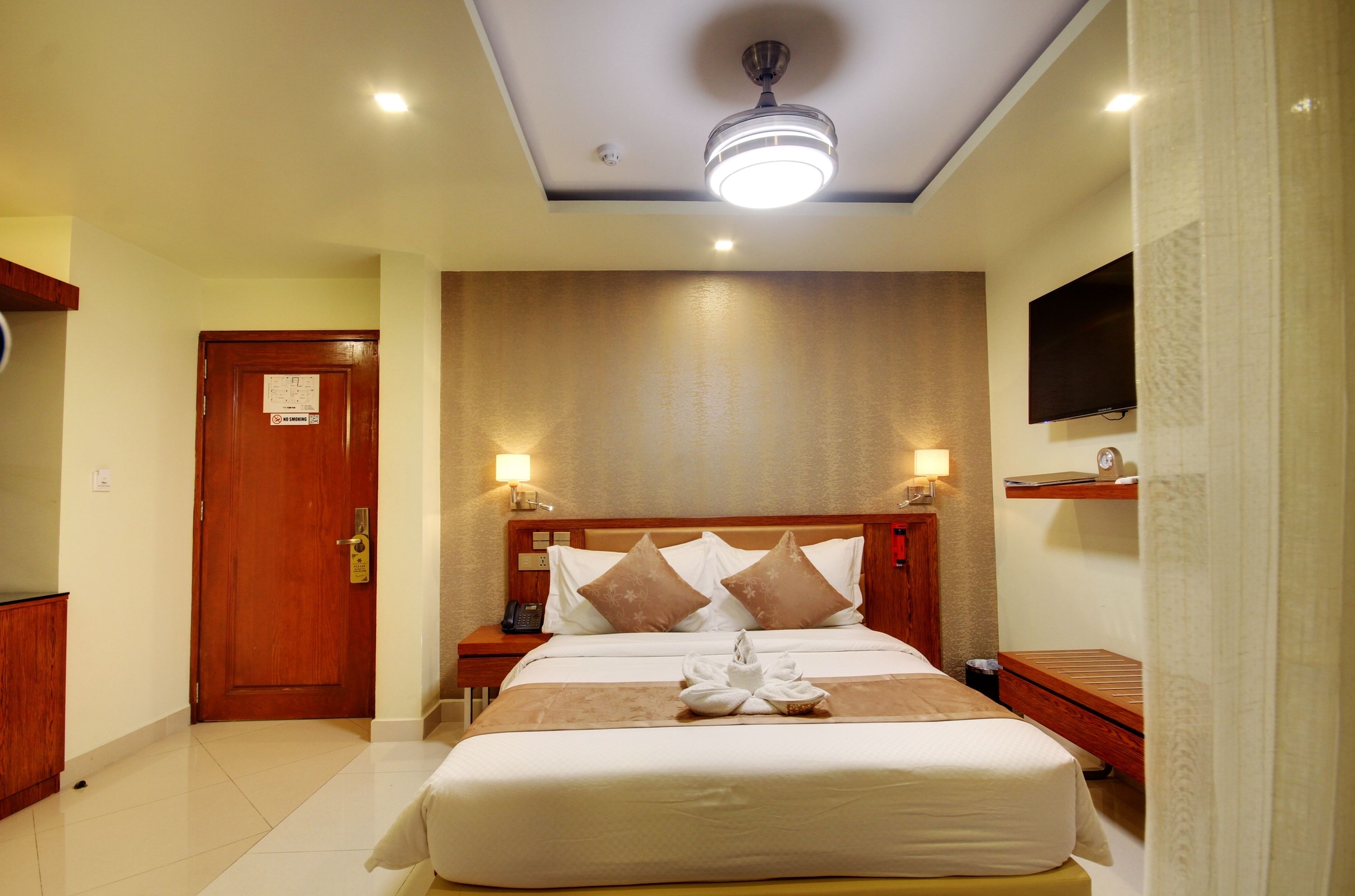 A hotel room with a large bed, two bedside tables, a ceiling fan, a flat-screen TV, and a wooden nightstand.