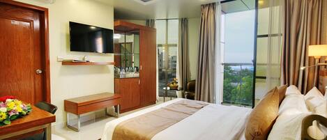 A hotel room with a bed, a desk with a chair, a TV mounted on the wall, a wardrobe, and a window with curtains.
