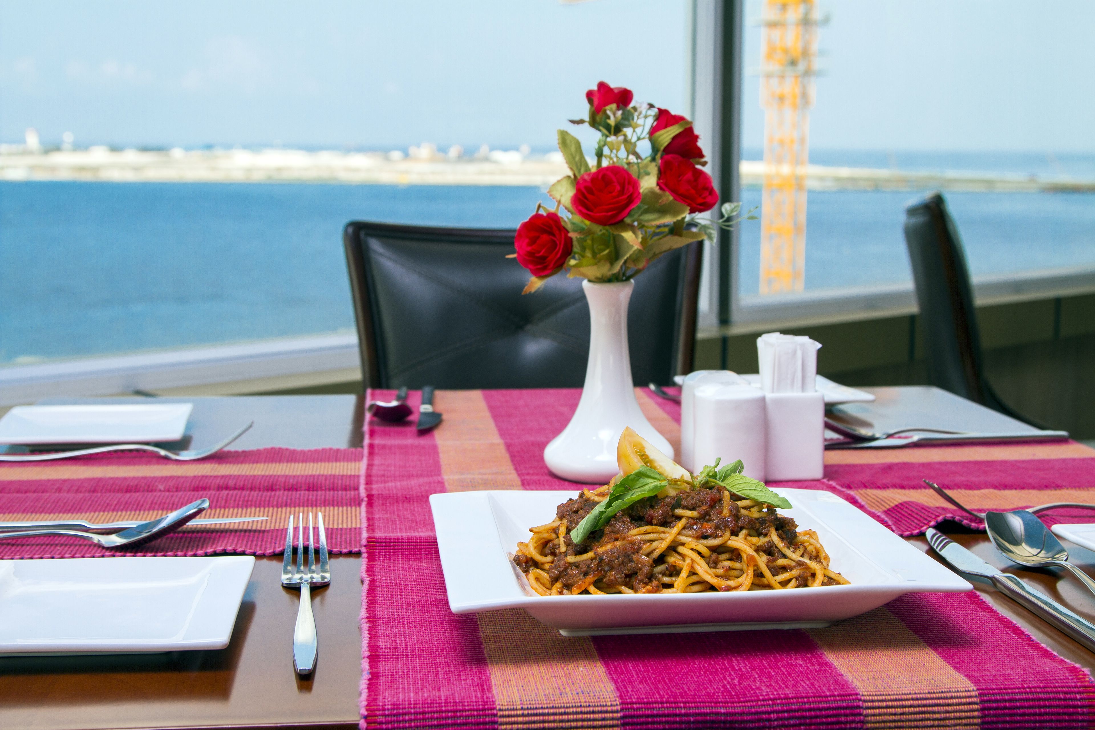A dining table with a plate of spaghetti, a vase of red roses, and a view of the sea.
