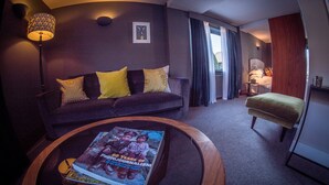 A modern living room with a dark-colored sofa, yellow cushions, a round coffee table with a magazine, a floor lamp, and a view of a bedroom with a bed and a nightstand.