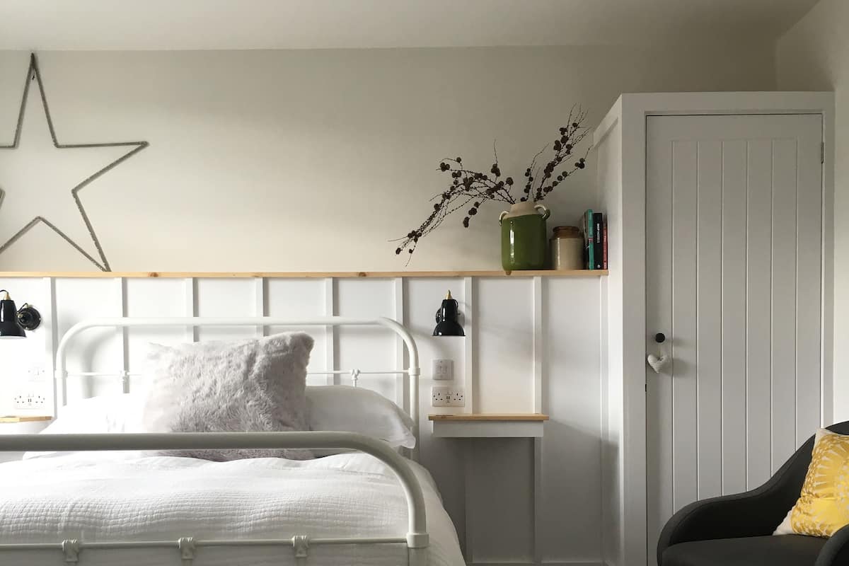 Un dormitorio con una cama de metal blanco, una mesita de noche, una silla, un estante con objetos decorativos y una puerta.