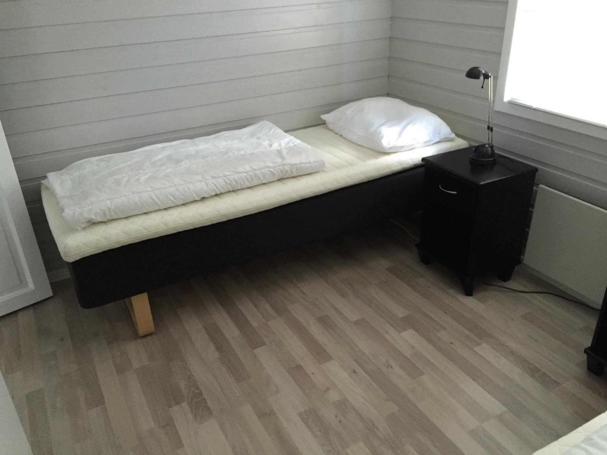 Una habitación pequeña con cama individual, piso de madera, una mesita de noche negra y una lámpara blanca.