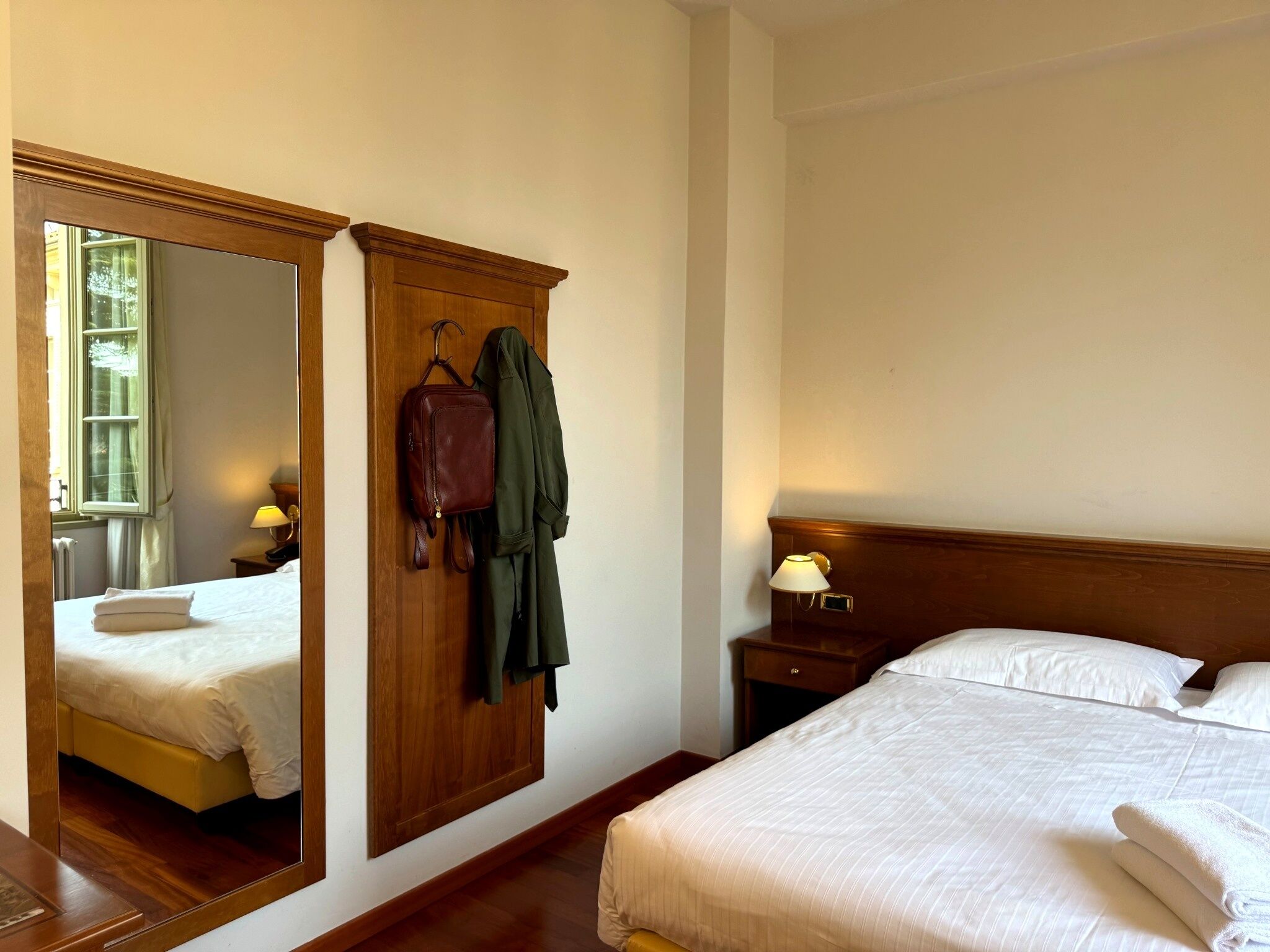 Ein Hotelzimmer mit großem Spiegel, einem Bett mit weißer Bettwäsche, einem Holzschrank und einem Nachttisch mit Lampe.