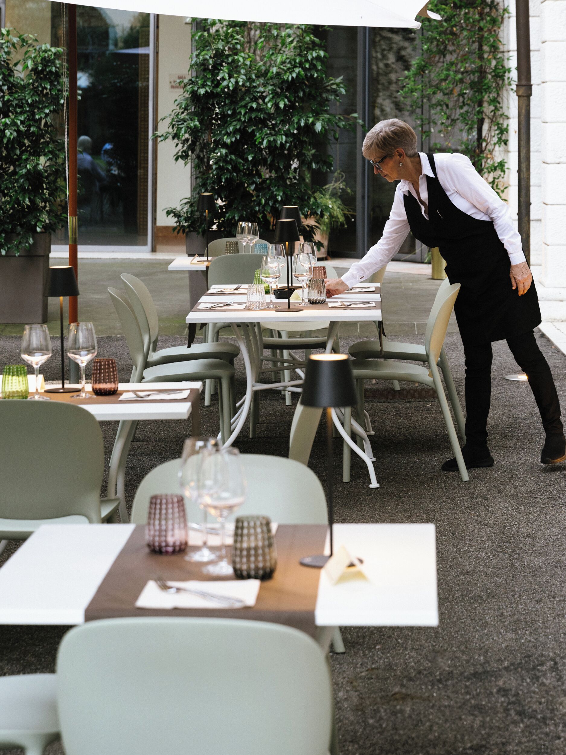 An outdoor dining area with tables set for two, each with a wine glass and cutlery. A waiter is attending to a table.