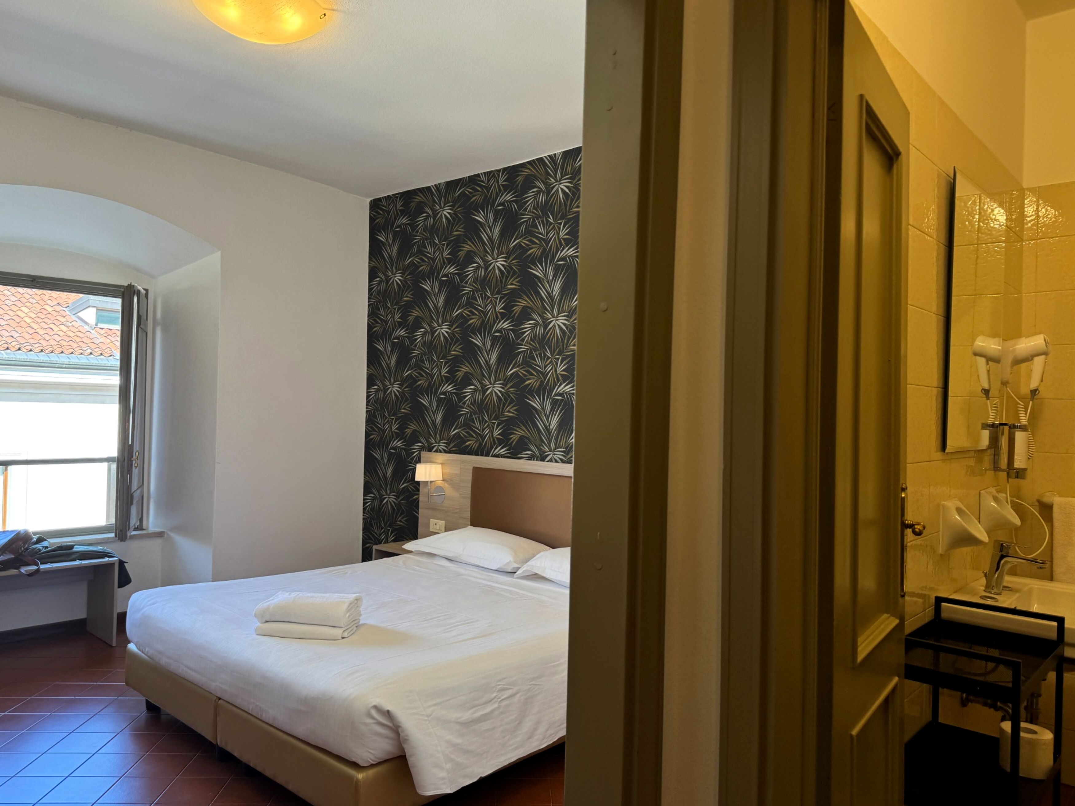A hotel room with a bed, a nightstand, a lamp, a bathroom with a sink, and a mirror.