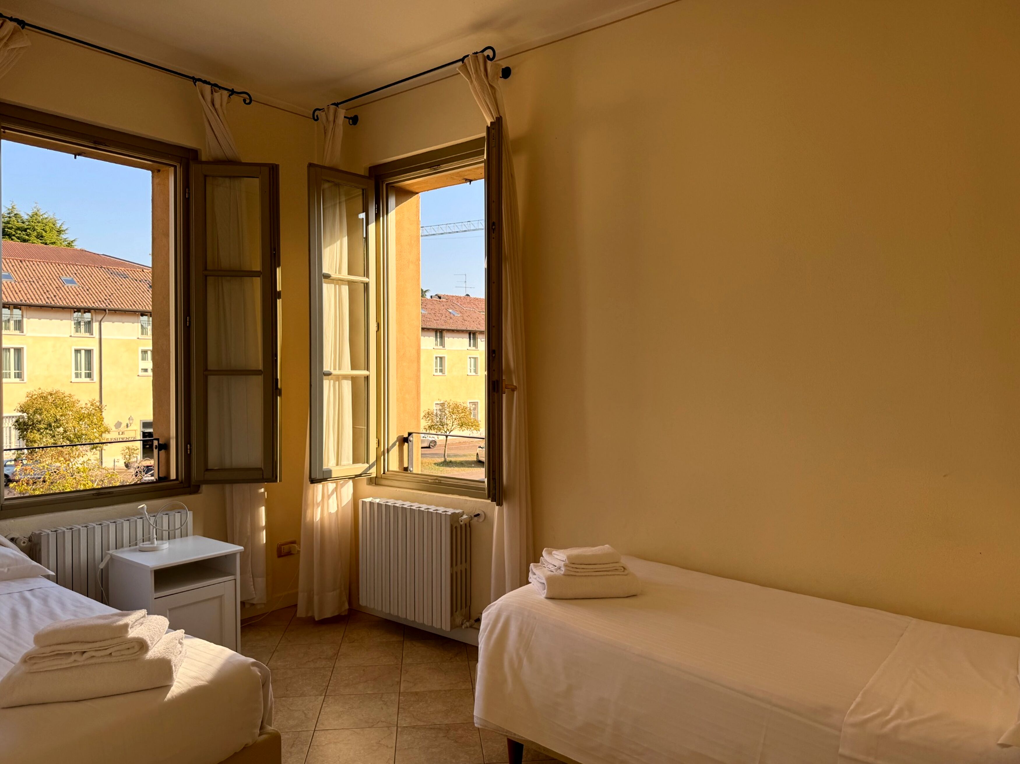 Habitación de hotel con dos camas, un radiador, una ventana con cortinas y vistas a edificios.