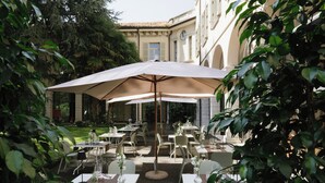 An outdoor dining area with tables and chairs, surrounded by greenery and a building with classical architecture. - Centro Paolo VI (Brescia)