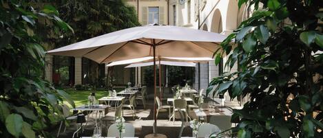 An outdoor dining area with tables and chairs, surrounded by greenery and a building with classical architecture.