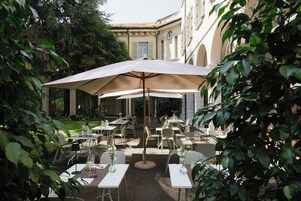 Un espace de restauration en plein air, avec des tables et des chaises, entouré de verdure et bordé d’un bâtiment à l’architecture classique.