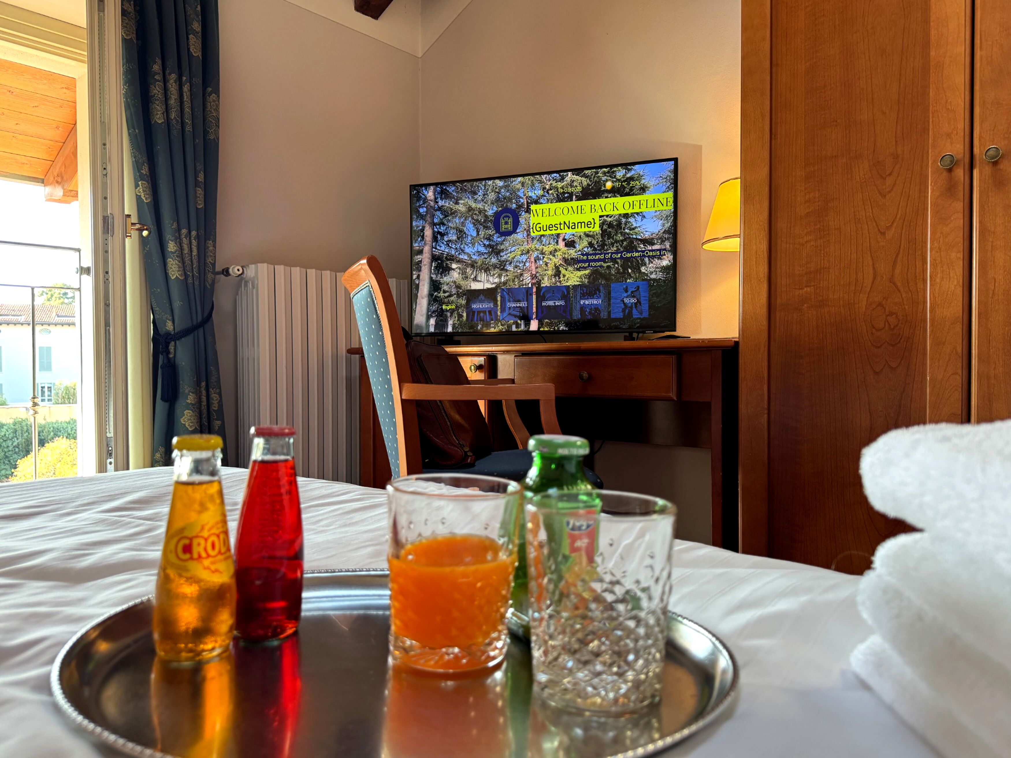 A hotel room with a bed, a television displaying a welcome message, a tray with drinks, and a view of the outdoors.