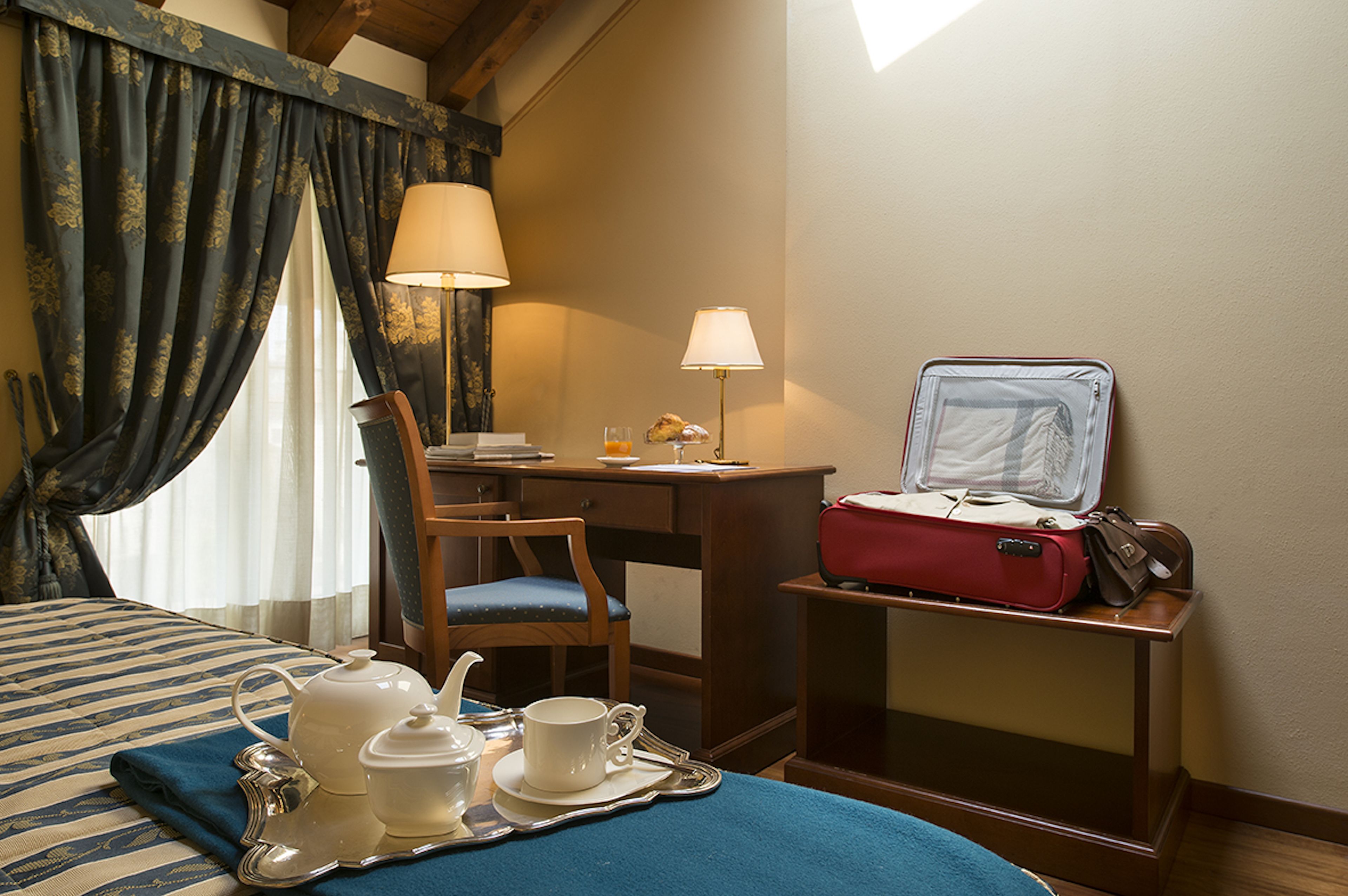 A hotel room with a bed, a desk, a chair, a table with a tray of tea and cups, a red suitcase, and a lamp.