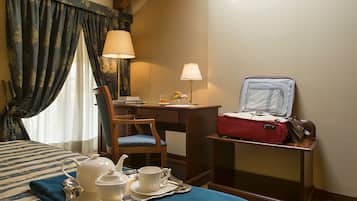 A hotel room with a bed, a desk, a chair, a table with a tray of tea and cups, a red suitcase, and a lamp.