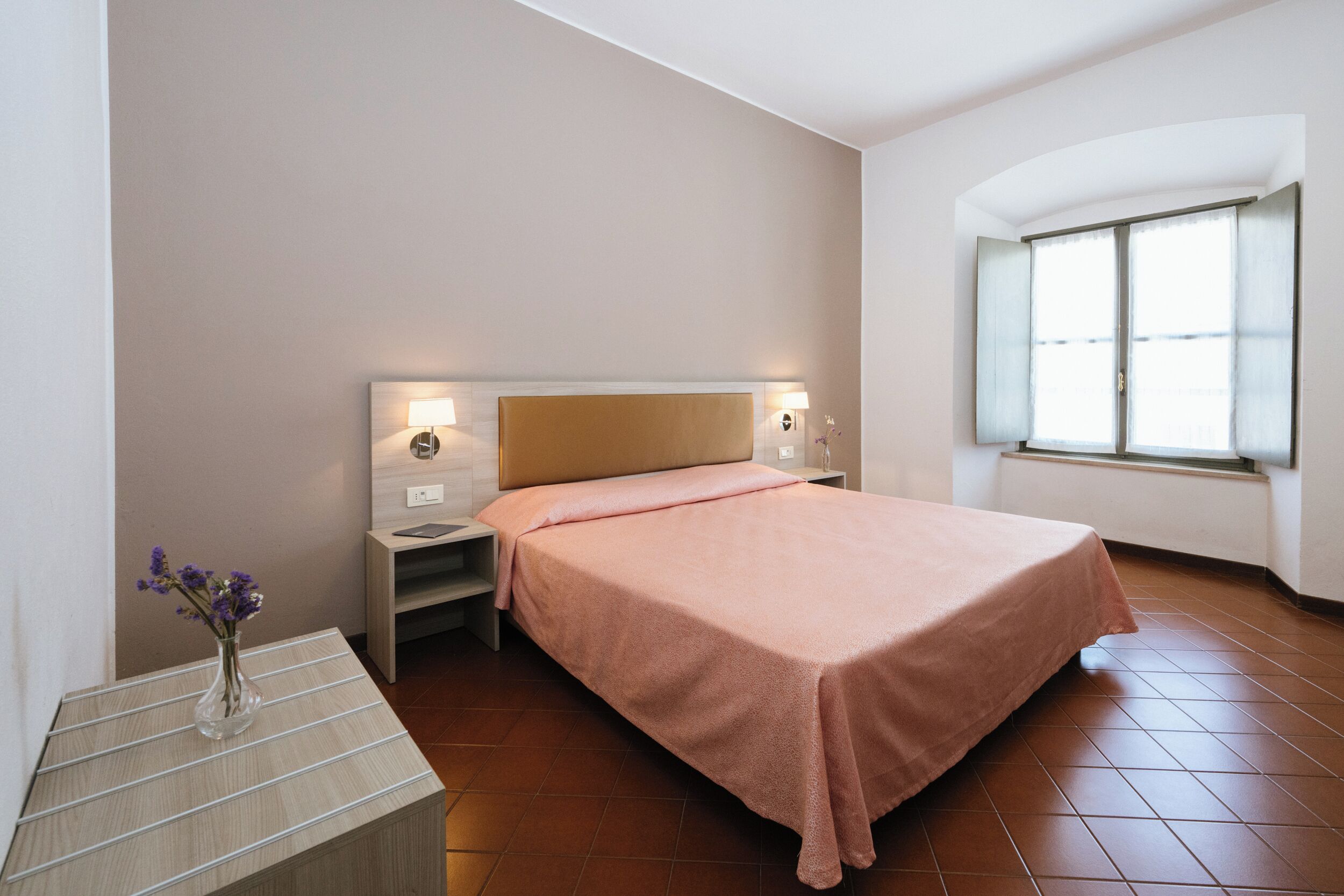 A hotel room with a bed, bedside tables, a vase with flowers, and a window with curtains.