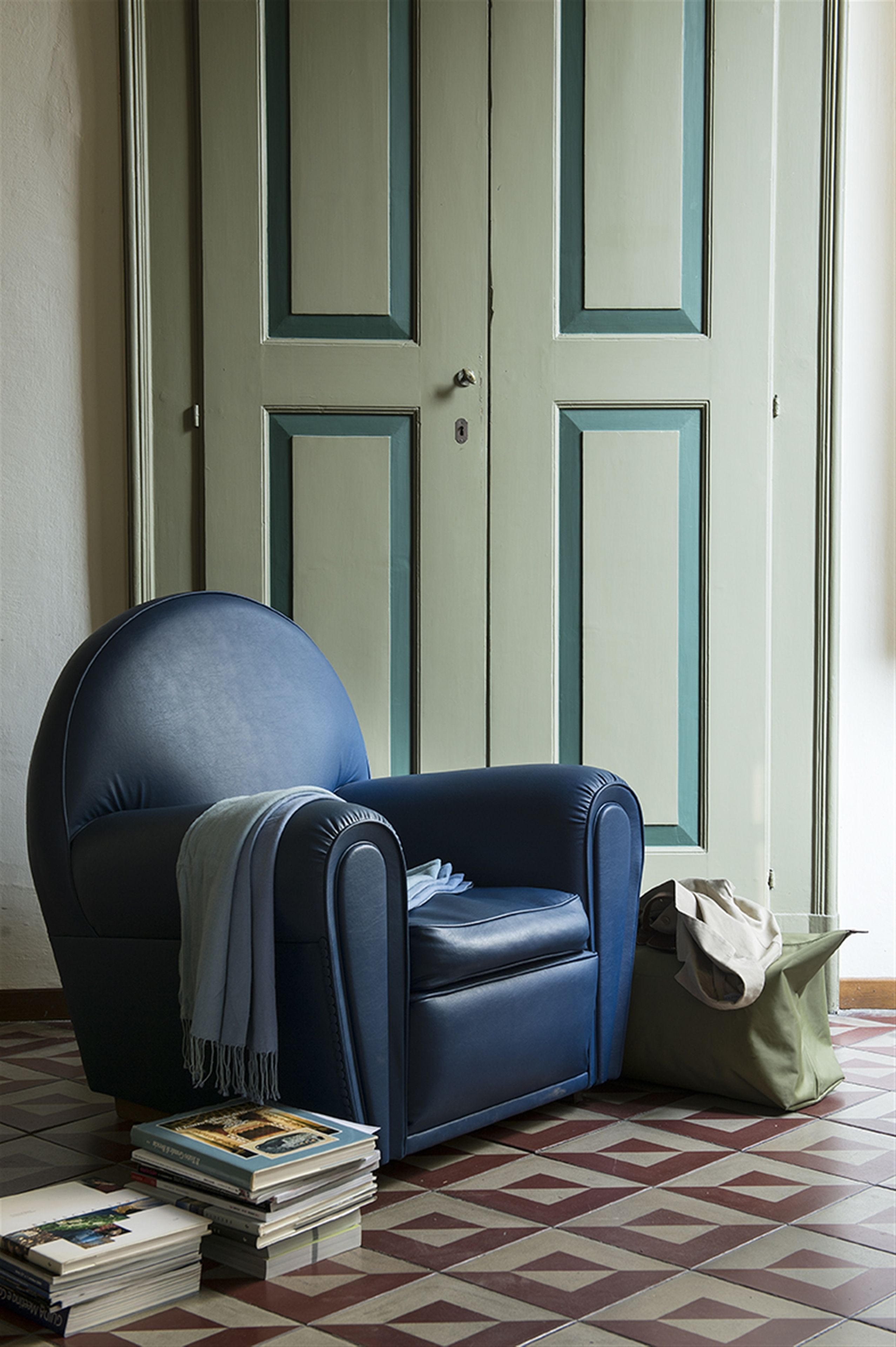Une pièce avec un fauteuil bleu, une pile de livres et un sac posé sur le sol.