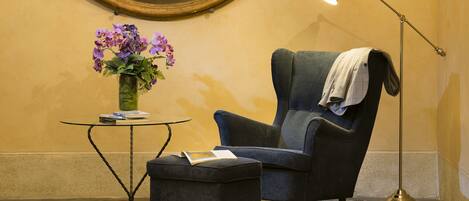 Una habitación con un sillón oscuro, una pequeña mesa redonda con un jarrón de flores, una lámpara de pie y un libro en el reposapiés.