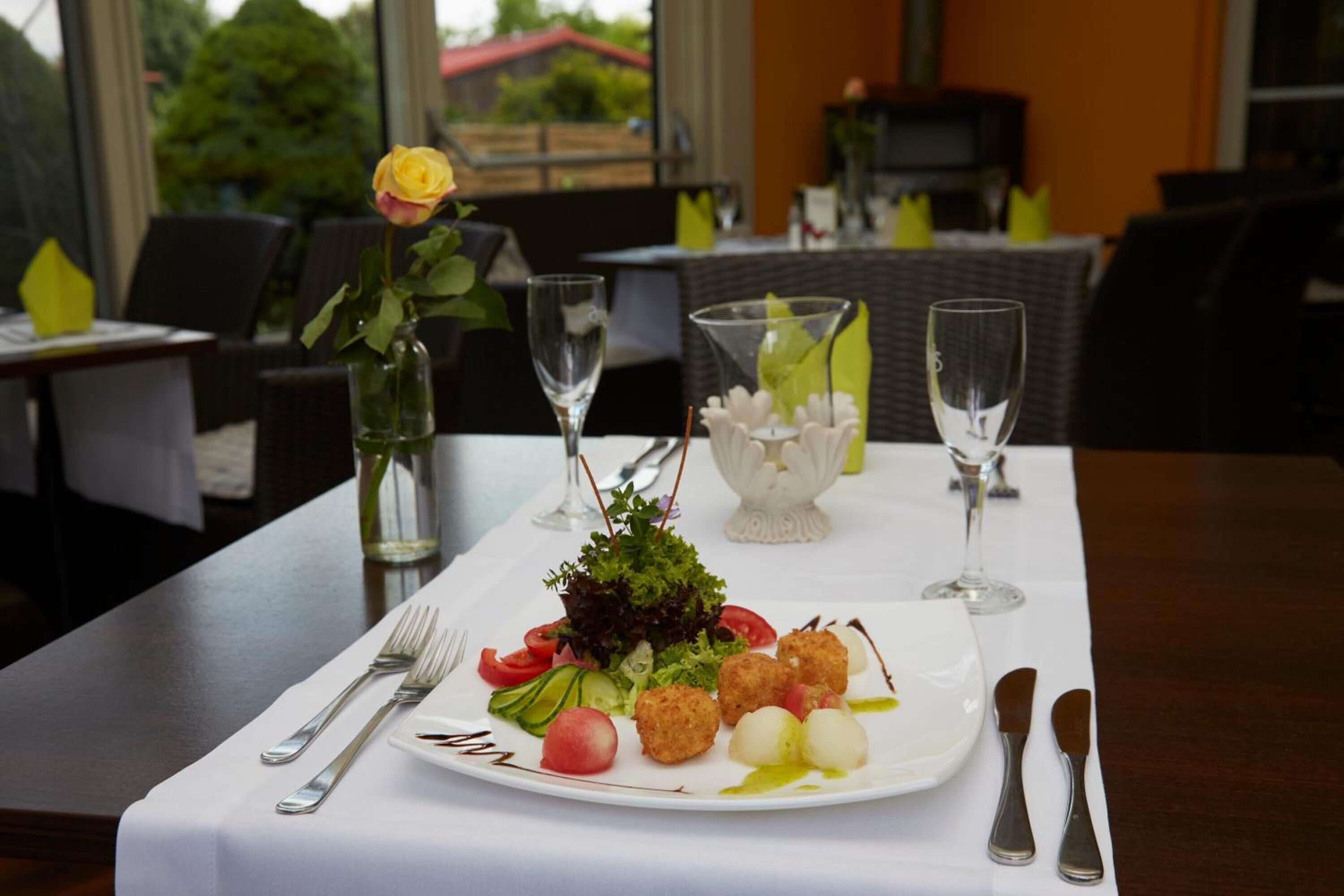 A dining table set with a plate of food, including a salad and croquettes, and a vase with a single yellow rose.