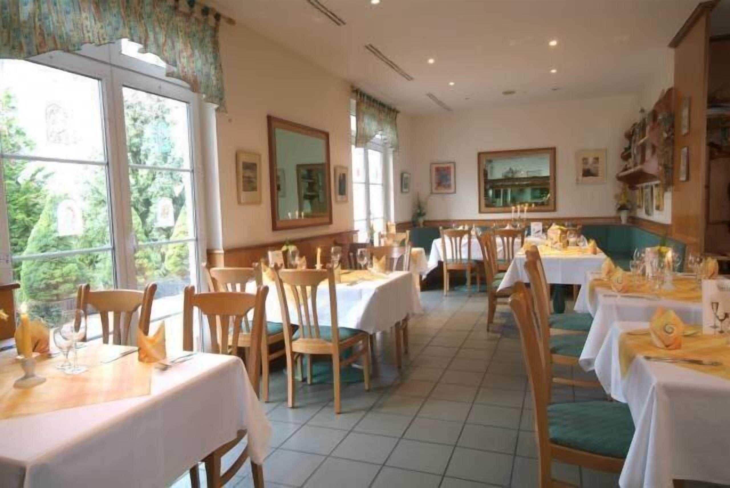 Un comedor con varias mesas dispuestas para comidas, ventanas amplias con cortinas y cuadros en las paredes.