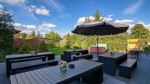 An outdoor patio with wooden tables and benches, a large umbrella, and a view of a garden and trees. - Flair Hotel Reuner (Zossen)