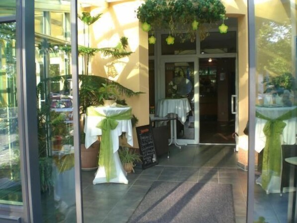 Una entrada con puertas de vidrio, una pequeña mesa con mantel blanco y adornos verdes, y plantas en macetas.