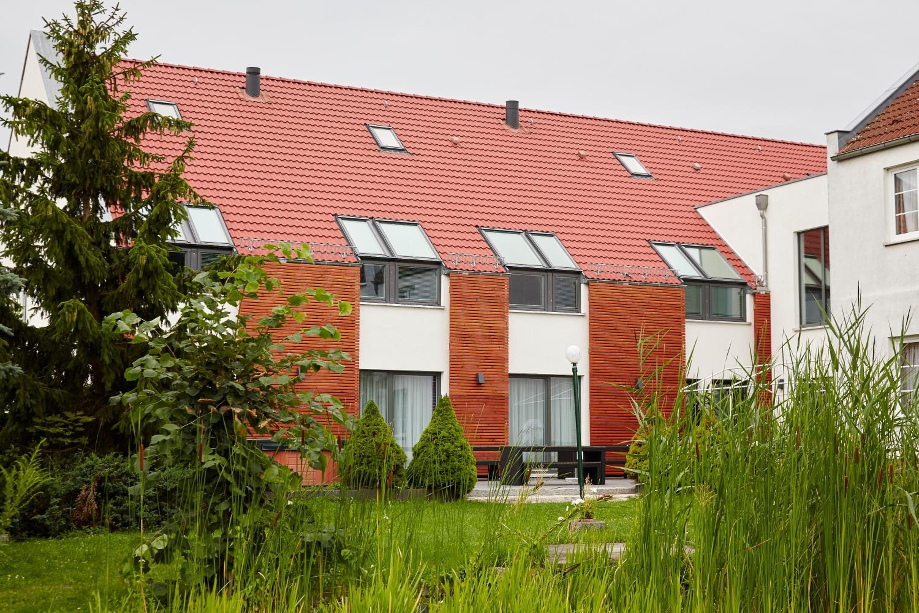 Un bâtiment moderne avec un toit recouvert de tuiles rouges, de nombreuses fenêtres et un jardin agrémenté de pelouse et d’arbres.