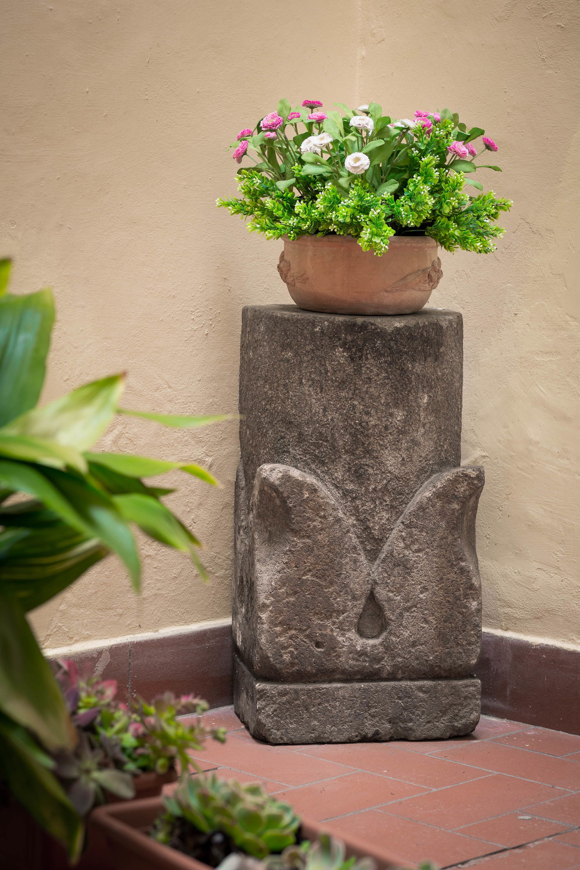 A stone planter with a flower arrangement on a tiled floor against a wall.