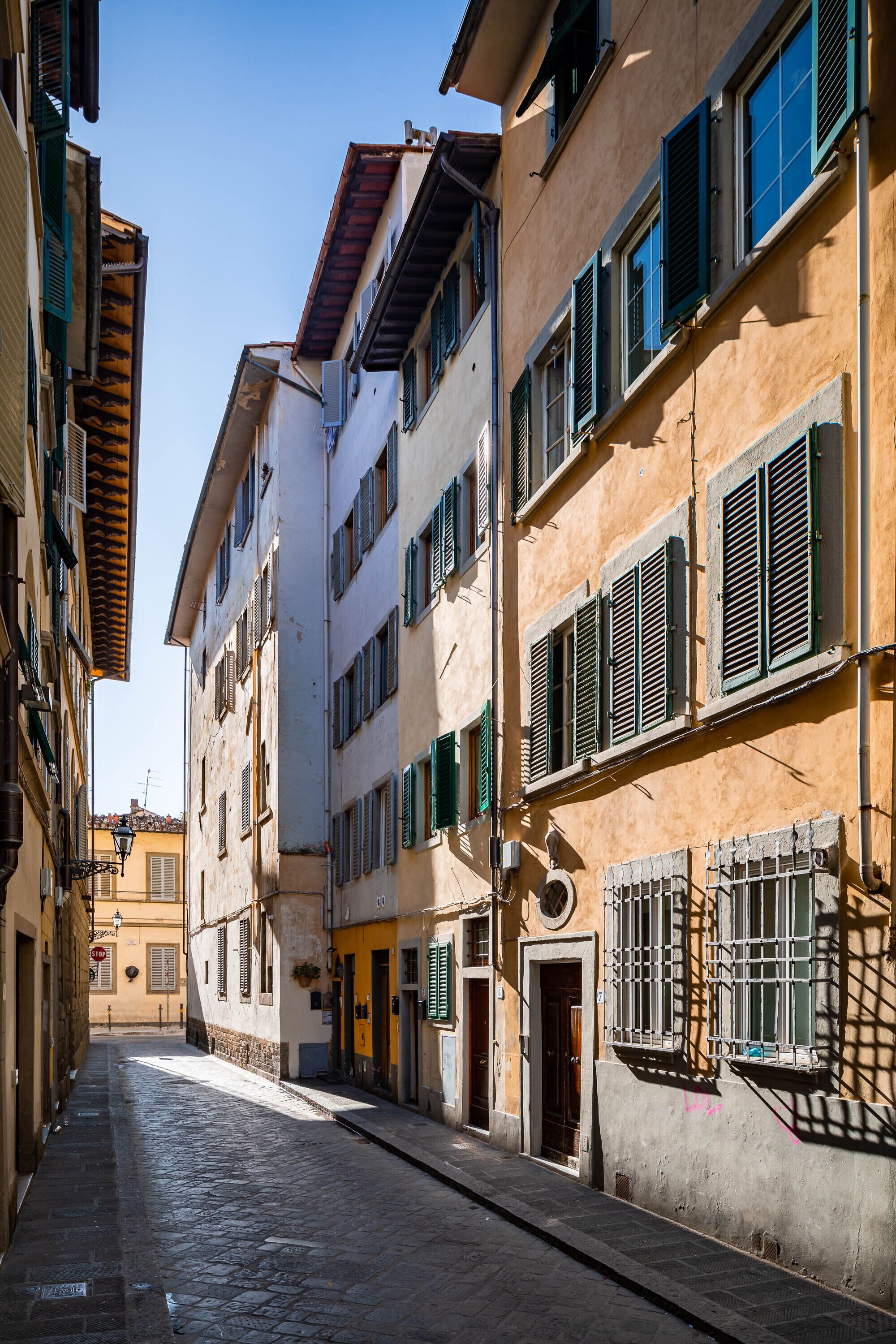 A narrow cobblestone street lined with multi-story buildings featuring traditional architecture, shutters, and a clear sky.