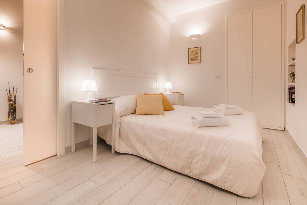 A modern bedroom with a large bed, white nightstands, and a wardrobe.