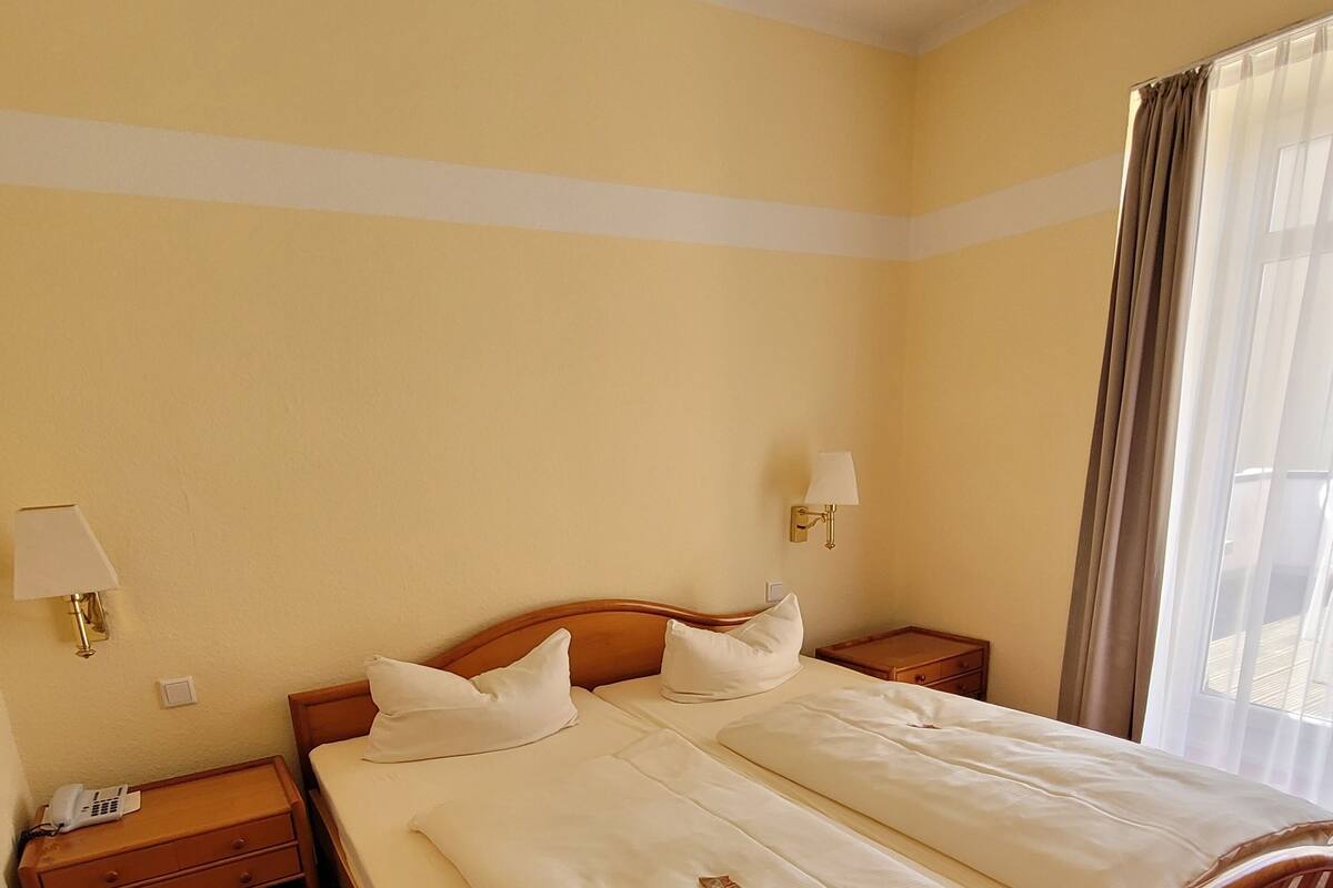 A double bed with a wooden frame, two bedside tables with lamps, a telephone on the left table, and a window with curtains on the right.