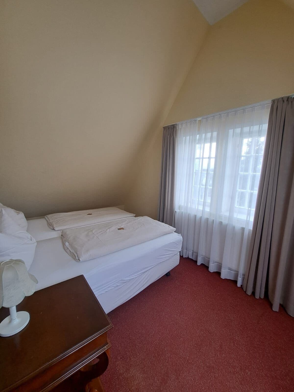 A hotel room with a bed, a wooden nightstand, and a window with curtains.