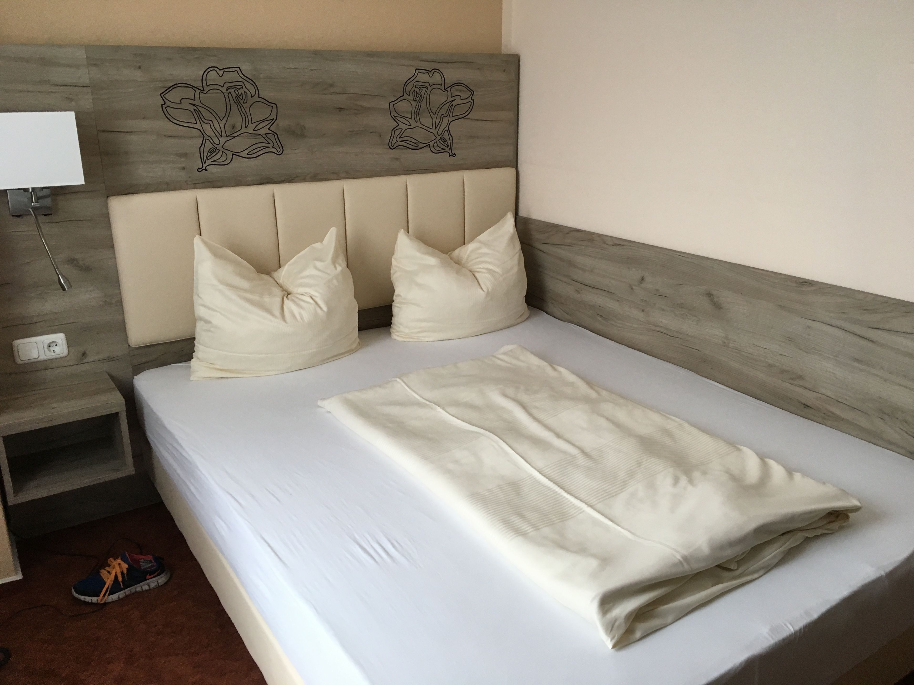 Een net opgemaakt bed met een opgevouwen plaid, twee kussens en een houten hoofdbord met bloemenmotieven.