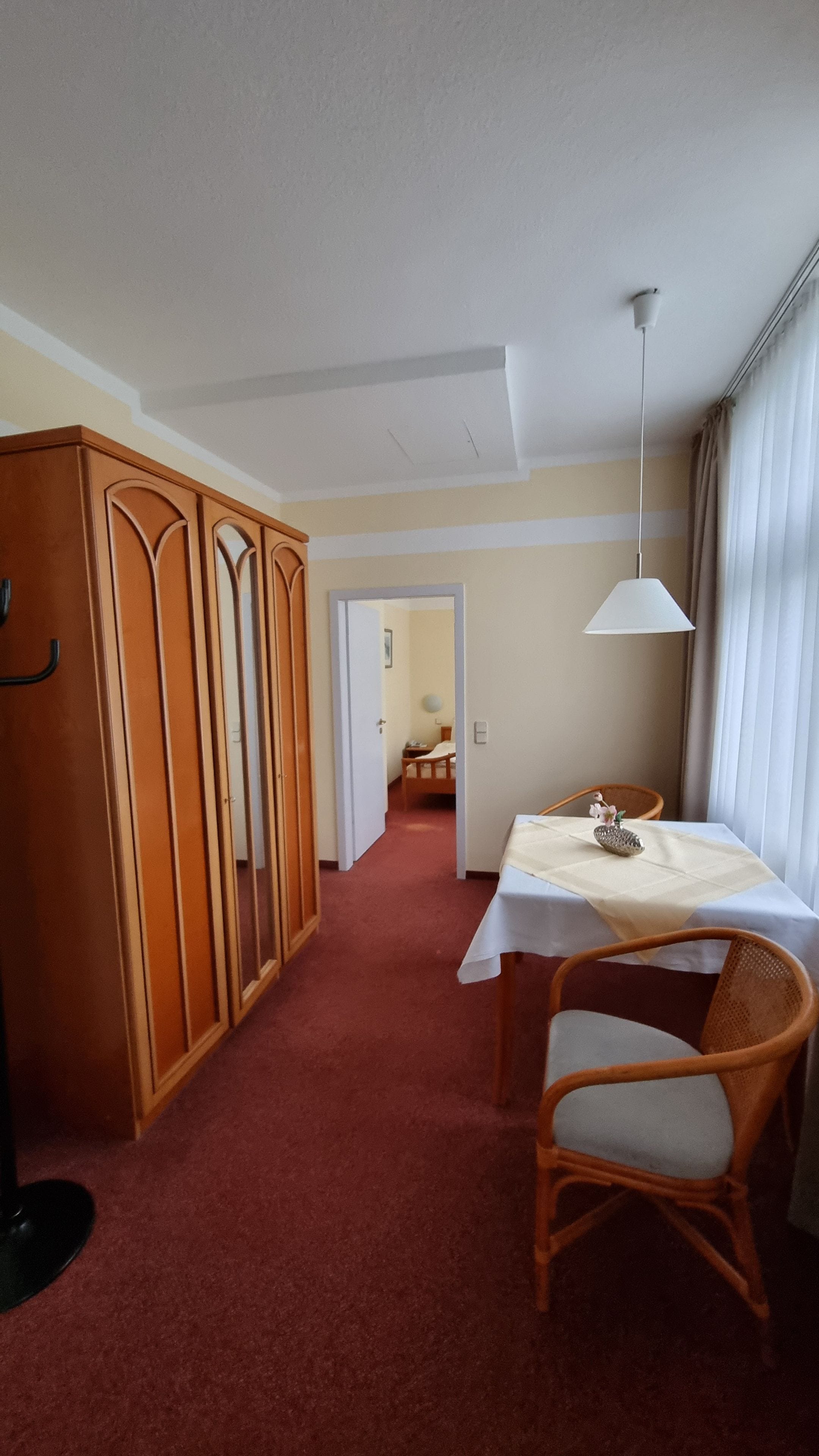 Habitació d'hotel amb una catifa vermella, un armari de fusta, una taula de dinar amb una cadira i una làmpada penjant.