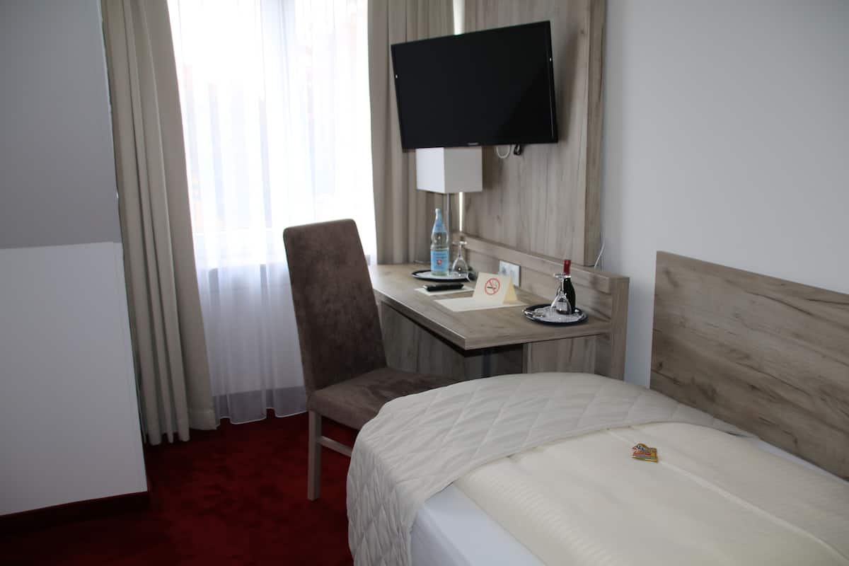 A hotel room with a bed, a desk with a chair, a television mounted on the wall, and a window with curtains.