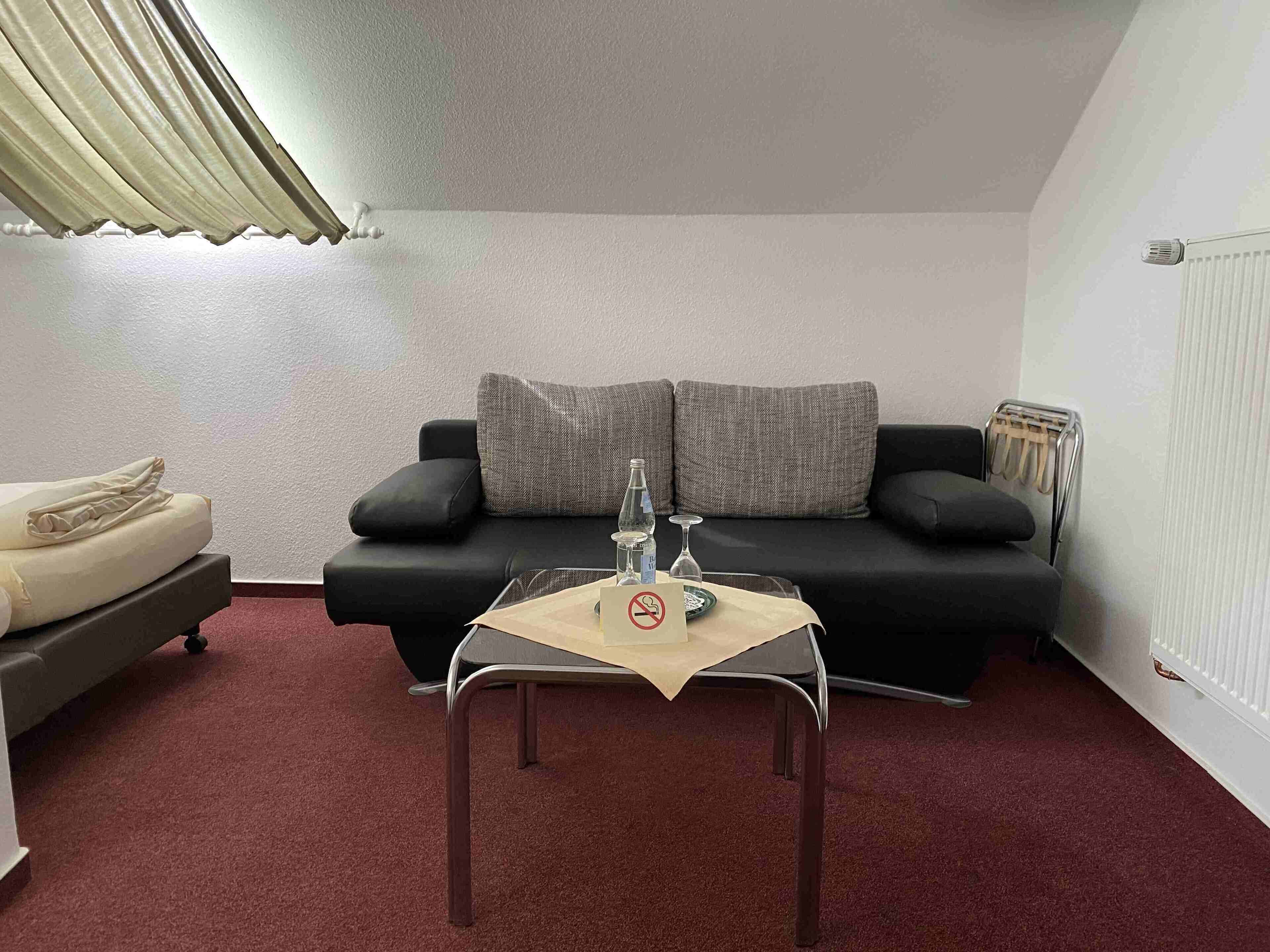 A small, cozy room with a black sofa, a small table with a bottle and glass, and a radiator.