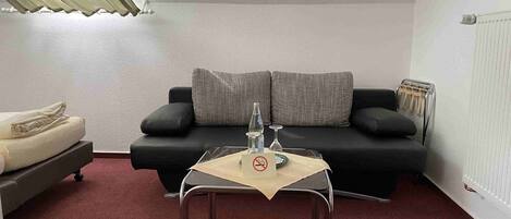 A small, cozy room with a black sofa, a small table with a bottle and glass, and a radiator.
