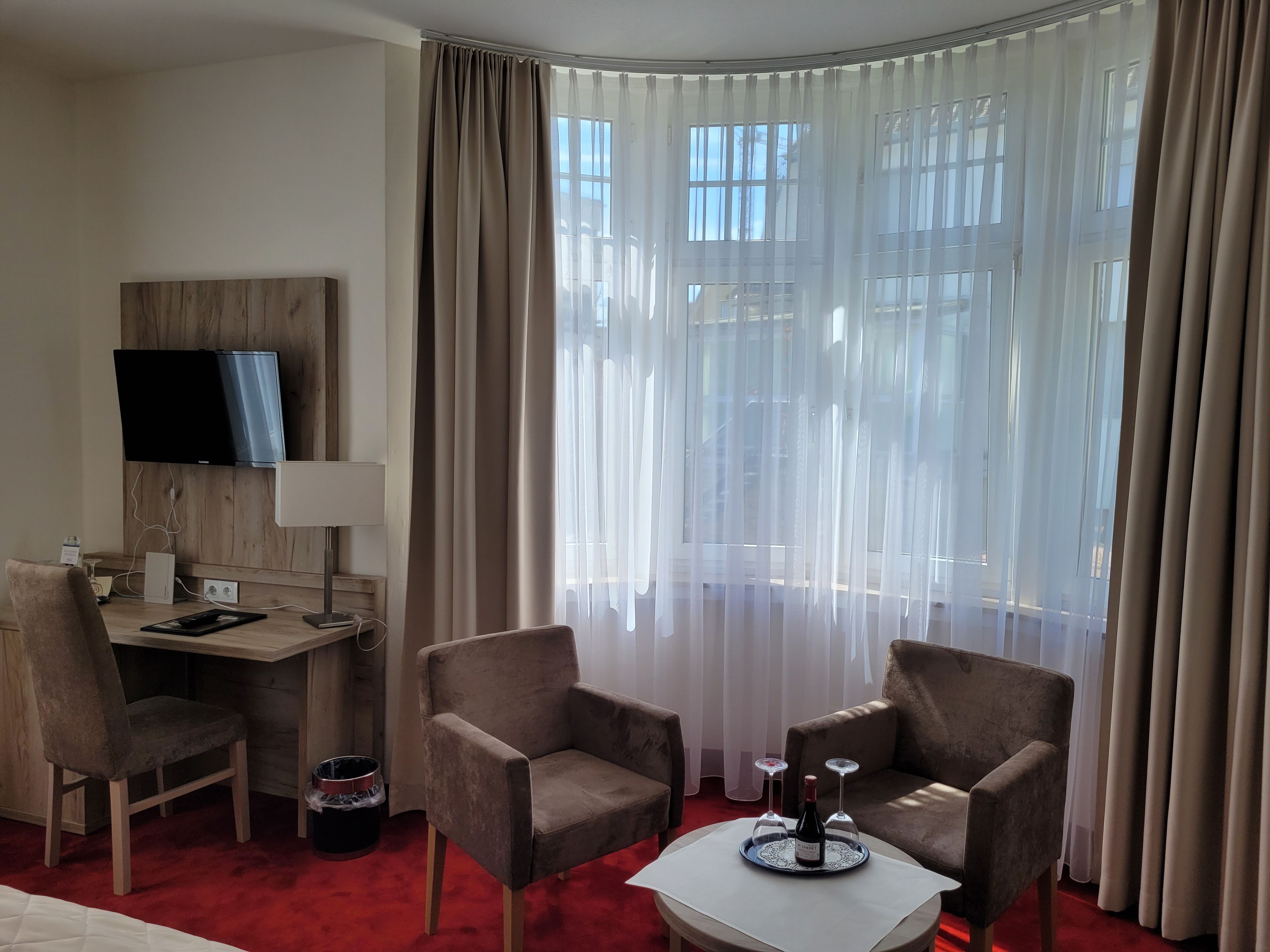 A hotel room with a desk, two armchairs, a TV mounted on the wall, and a large window with sheer curtains.