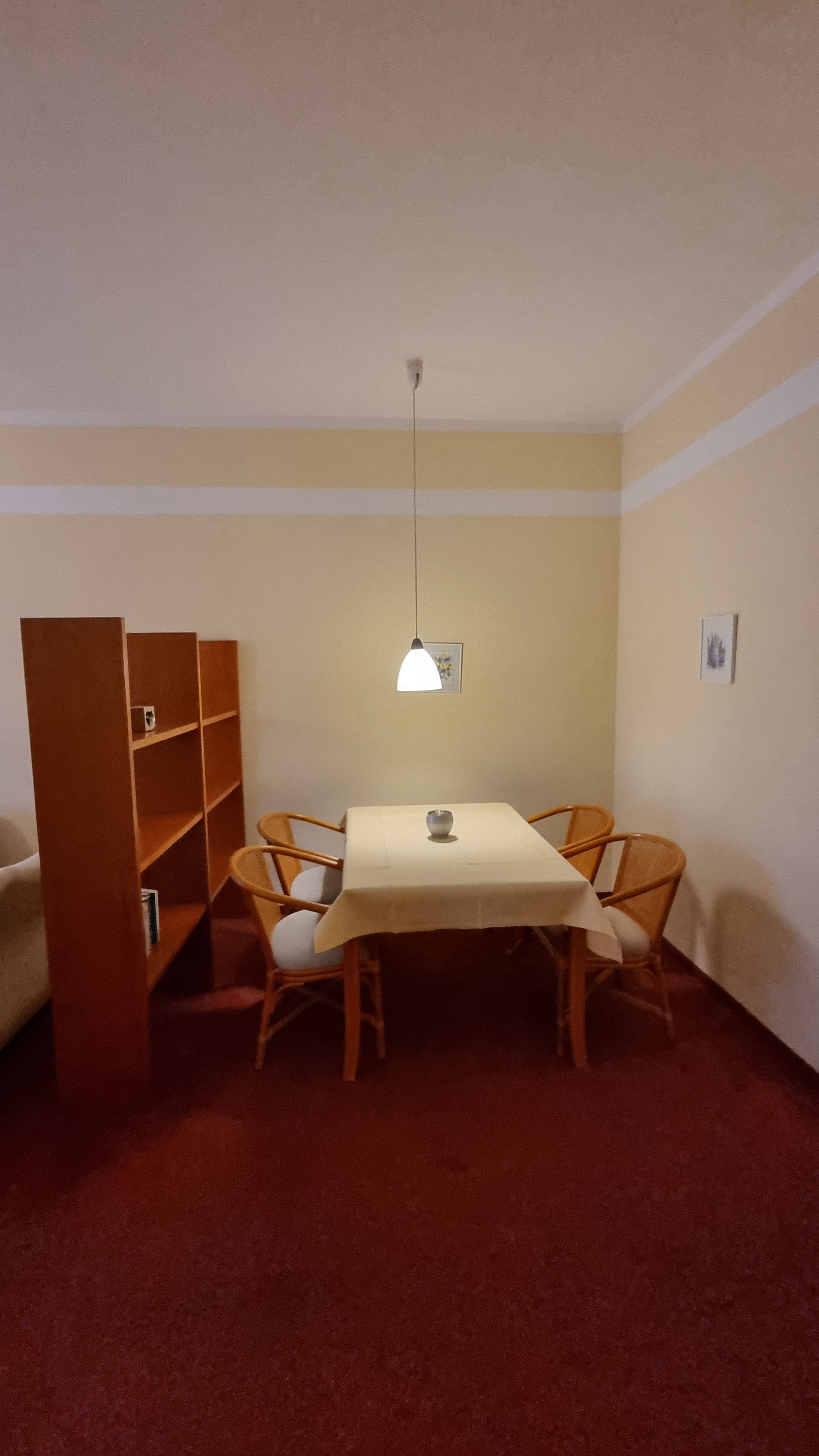 A room with a dining table, chairs, a bookshelf, and a hanging light fixture.