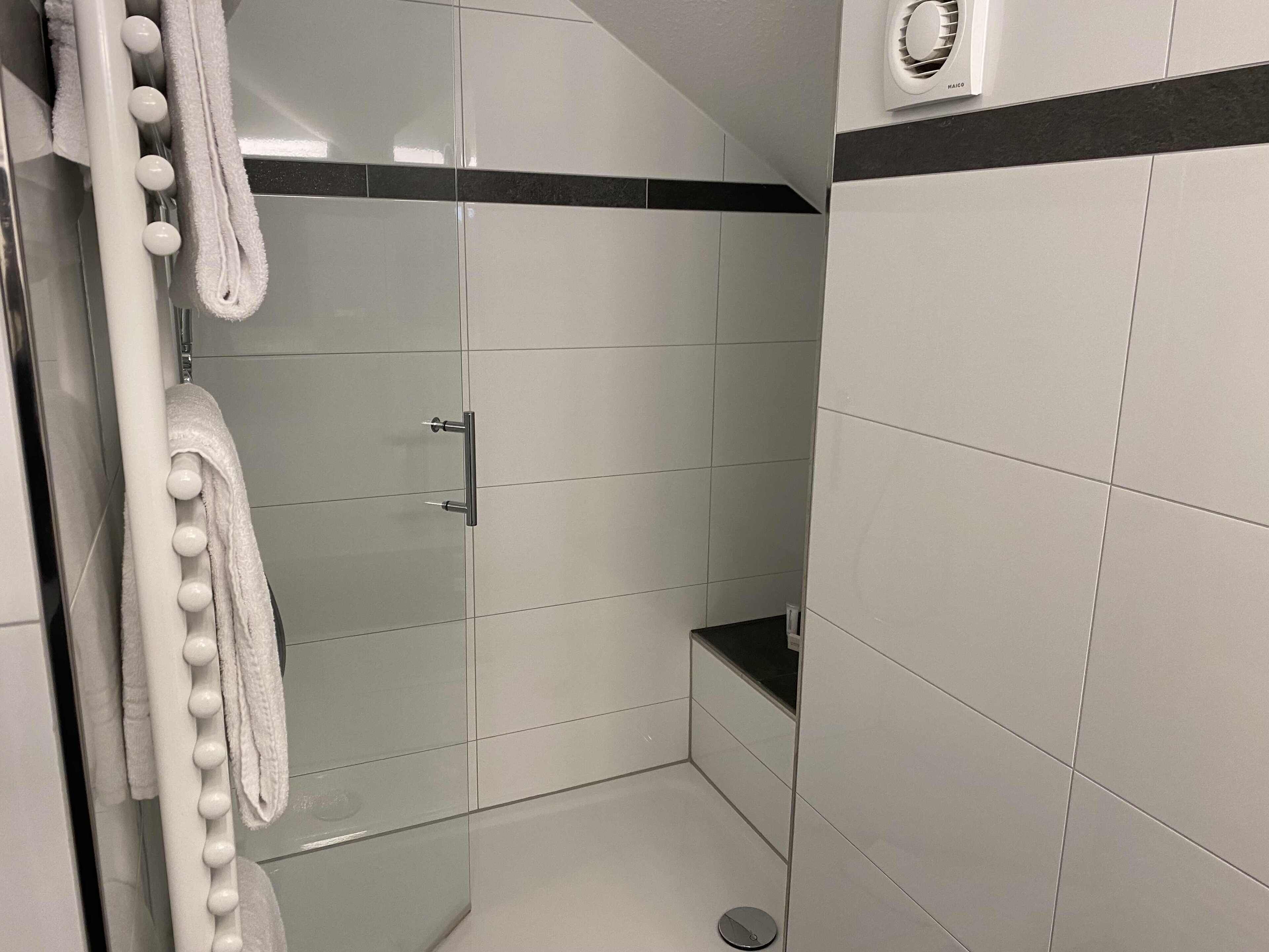 Un baño moderno con azulejos blancos, mampara de vidrio y toallero con toallas blancas.