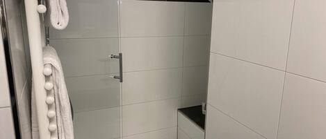 Un baño moderno con azulejos blancos, mampara de vidrio y toallero con toallas blancas.