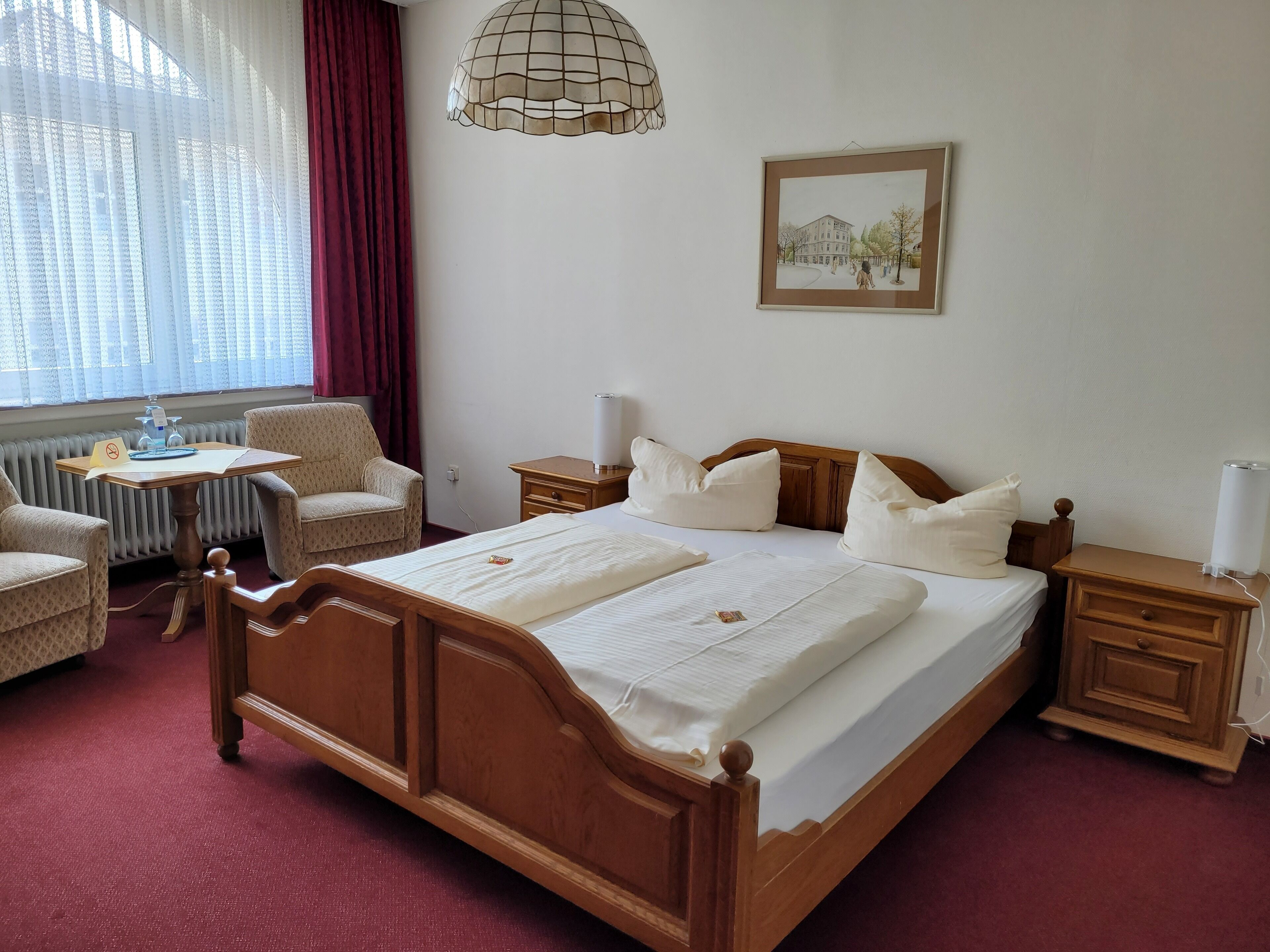 A hotel room with a wooden bed, a small table with a chair, a radiator, a lamp, and a framed picture on the wall.