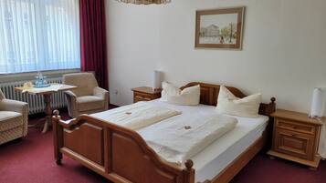 A hotel room with a wooden bed, a small table with a chair, a radiator, a lamp, and a framed picture on the wall.