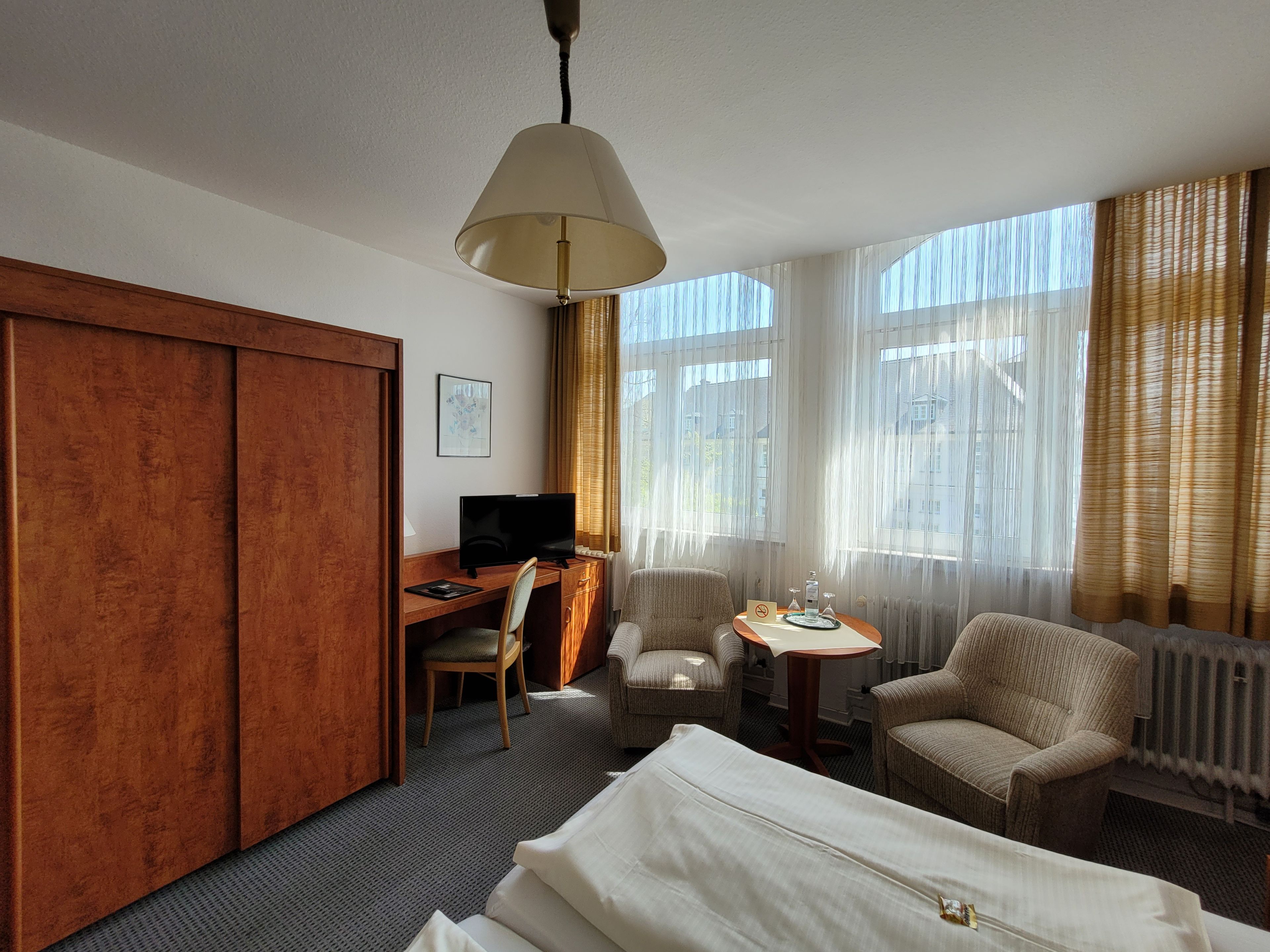 Ein Hotelzimmer mit einem Bett, einem Schreibtisch mit Computer, einem Sessel, einem kleinen Tisch mit Lampe und großen Fenstern mit Vorhängen.