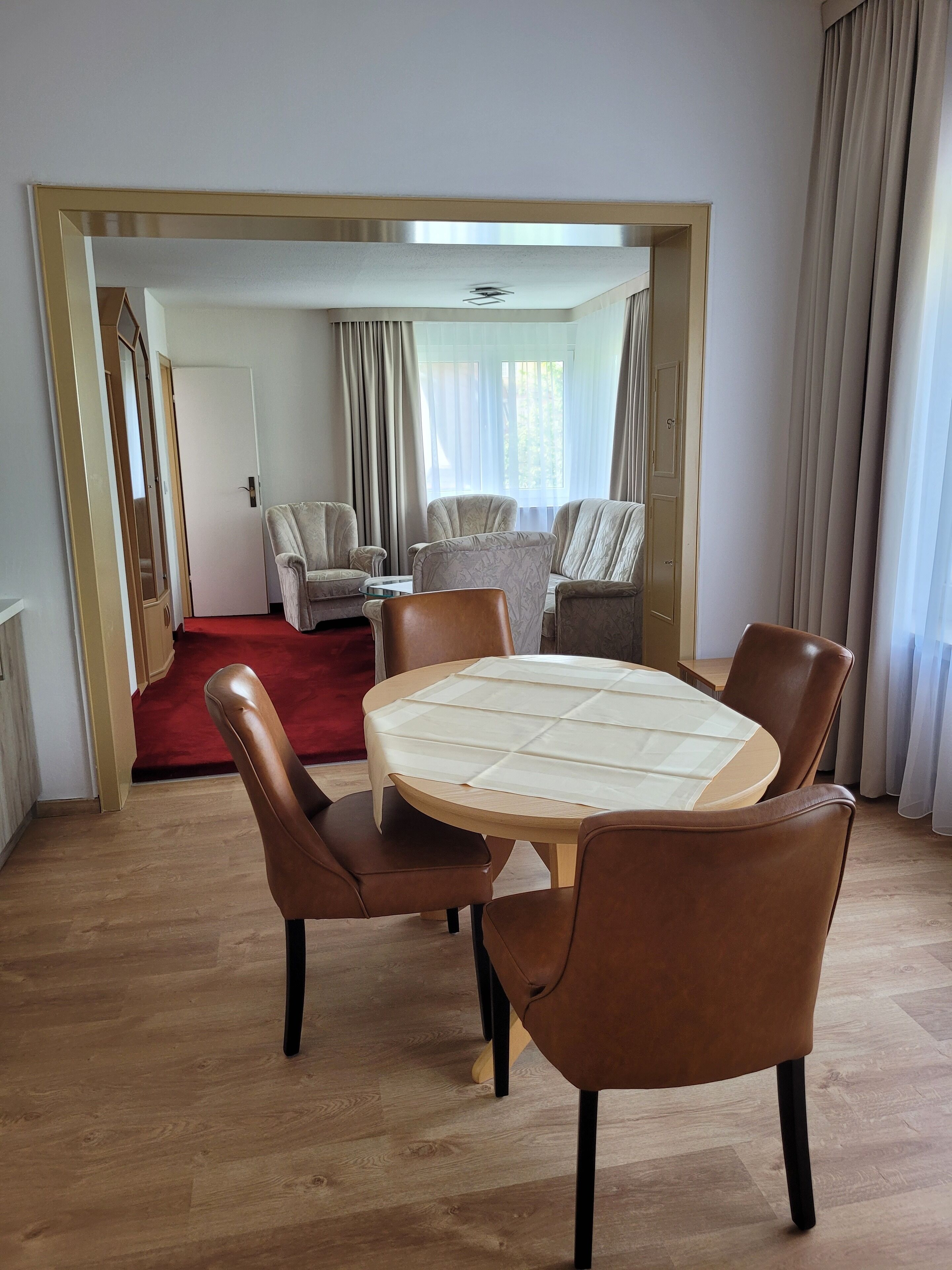 Una habitación con una mesa redonda, cuatro sillas, una alfombra roja y vistas a una sala de estar con sofá y cortinas.