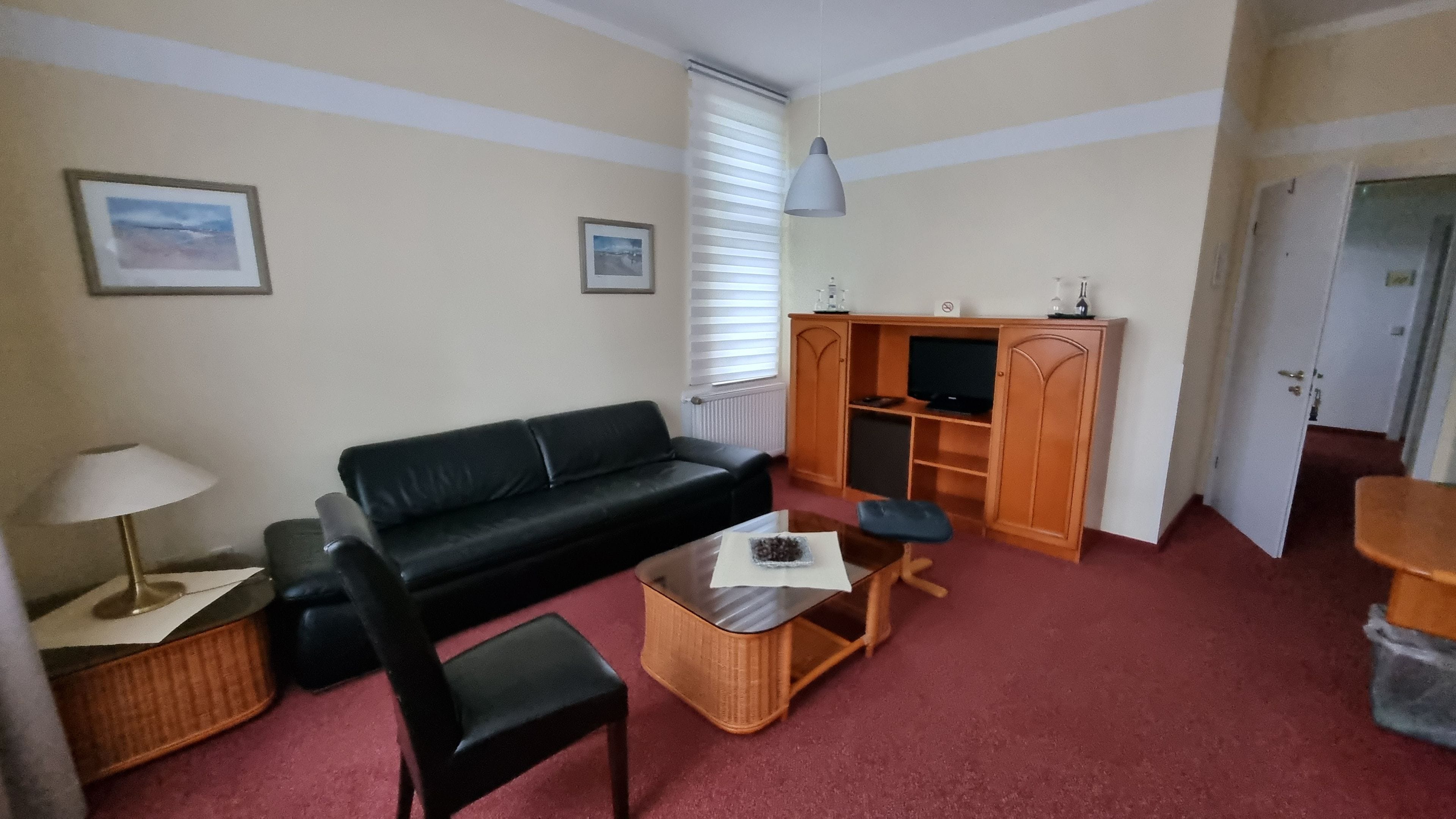 Een woonkamer met een zwarte bank, een houten salontafel, een zwarte fauteuil, een lamp, een televisie op een houten kast en een deur naar een andere kamer.