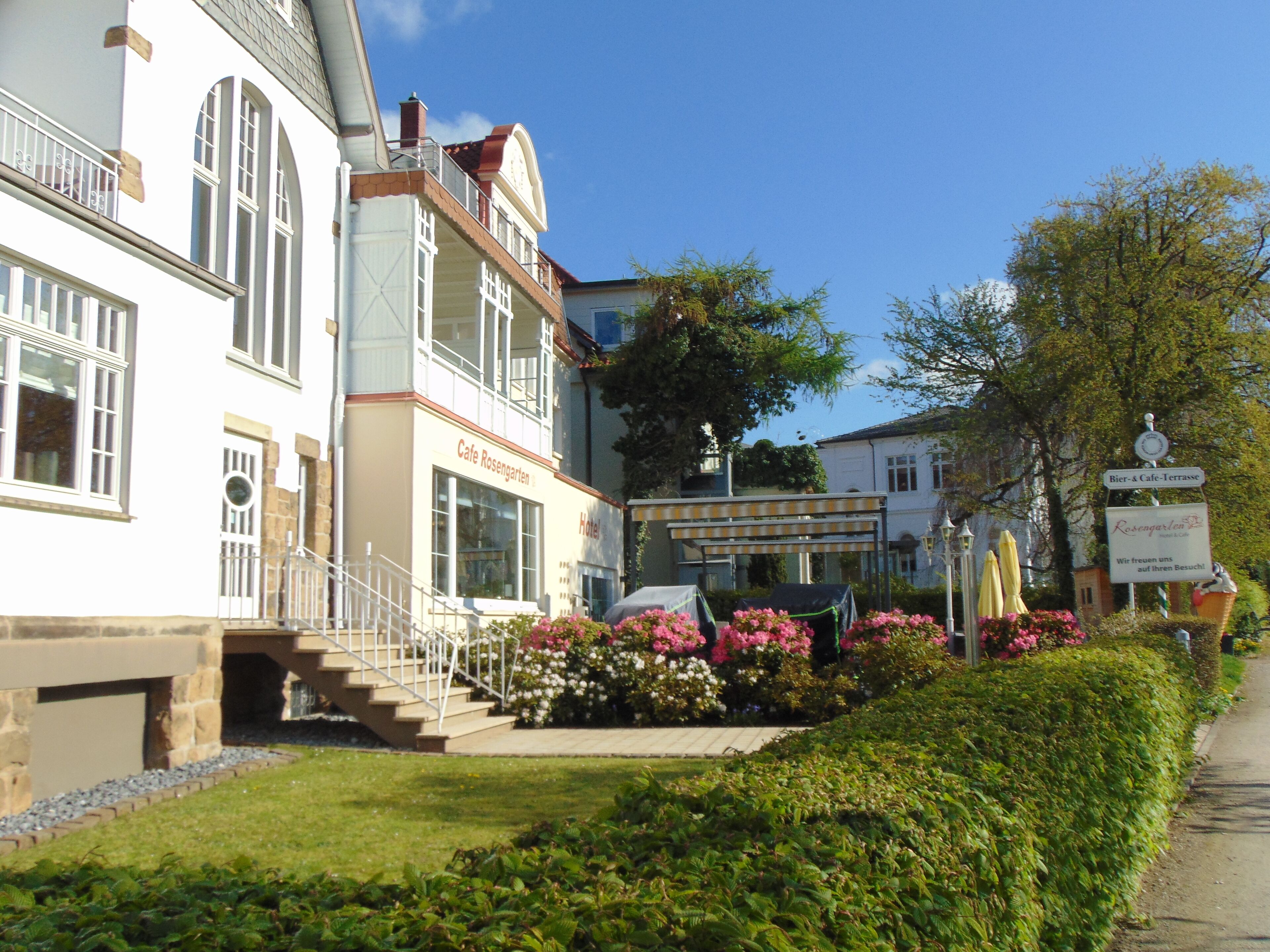 Un edificio de dos pisos con un café y letrero de hotel, una escalera que conduce a la entrada, y un jardín con flores y arbustos.