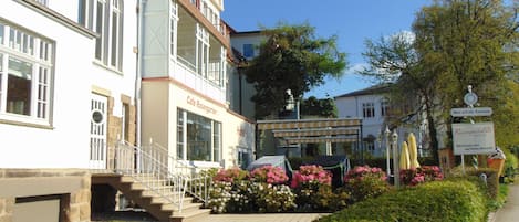 Un edificio de dos pisos con un café y letrero de hotel, una escalera que conduce a la entrada, y un jardín con flores y arbustos.